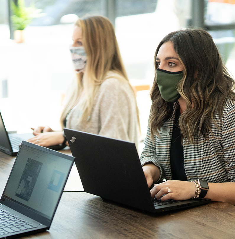two women wearing face masks sitting at table using laptops